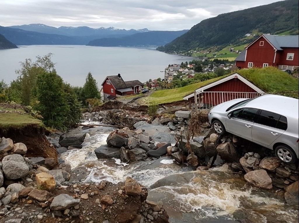 Flooding river down a steep hill, car on the edge. Red houses in the background.