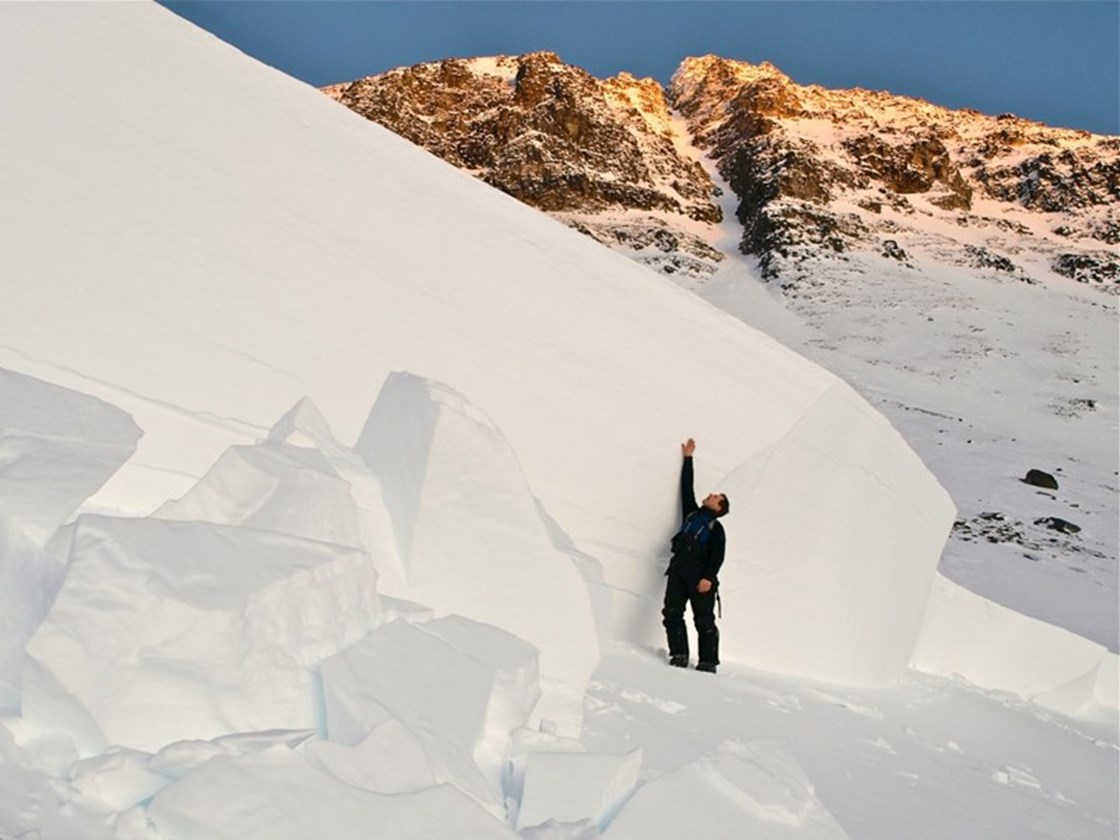 Et nærbilde av bruddkanten til et ekstremt stort snøskred. For målestokk står det en mann med oppstrakt hånd på siden og det er fremdeles nesten 2 meter til toppen av bruddkanten.