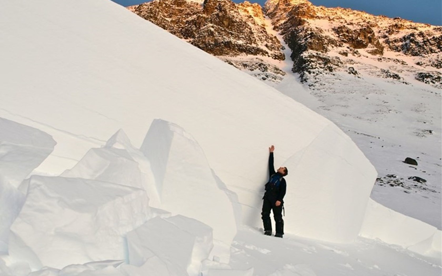 Et nærbilde av bruddkanten til et ekstremt stort snøskred. For målestokk står det en mann med oppstrakt hånd på siden og det er fremdeles nesten 2 meter til toppen av bruddkanten.